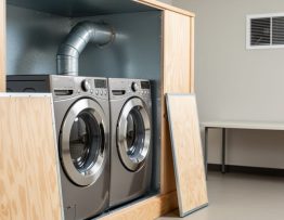 Commercial front-loading dryer in a custom fire-rated plywood cabinet with metal-lined interior; removable access panel set aside exposing rigid 4-inch galvanized duct with a single gentle bend; soft daylight; blurred background with makeup air vent and folding table; no text or logos.