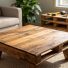 Handcrafted coffee table made from reclaimed pallet boards with visible grain, knots, and nail holes, photographed at a three-quarter angle in a bright living room with a neutral sofa, rug, houseplant, and softly blurred stack of pallets and hand tools in the background.
