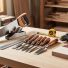 Essential beginner woodworking tools—handsaw, square, block plane, chisels, hammer, tape measure, drill, and sandpaper—arranged on a workbench with a small piece of wood.